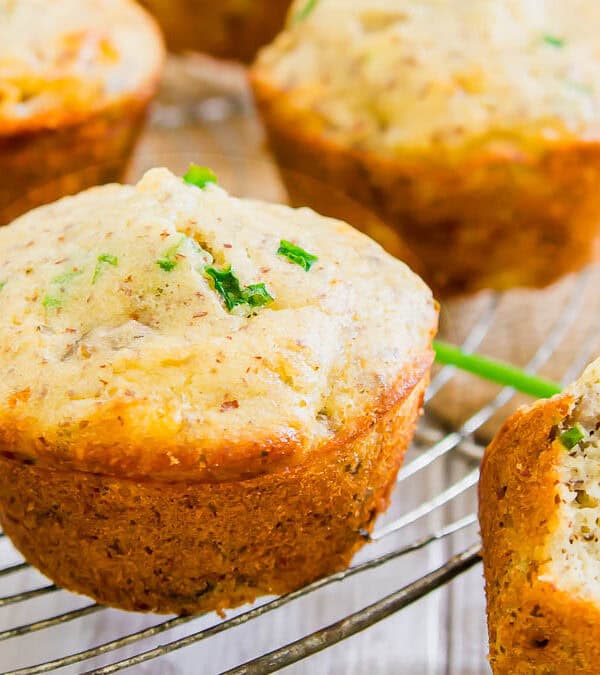 Close-up of cheddar sausage muffins cooling on a wire rack, with herbs sprinkled on top. One muffin is partially cut open, revealing a fluffy interior.
