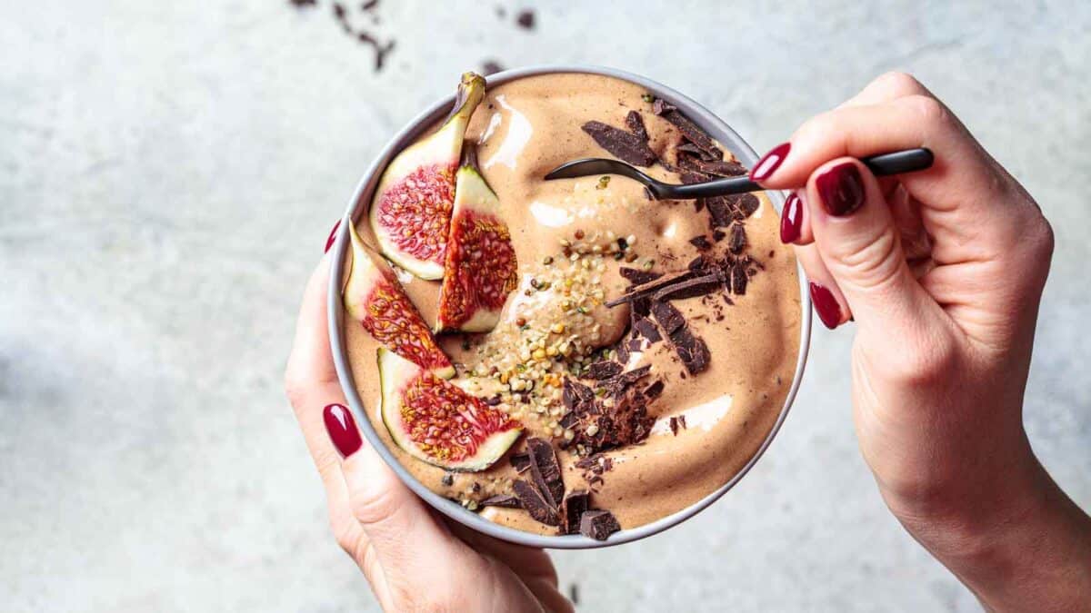 A person with red-painted nails holds a bowl of chocolate smoothie topped with sliced figs, chocolate shavings, and seeds, using a spoon to scoop from the bowl.