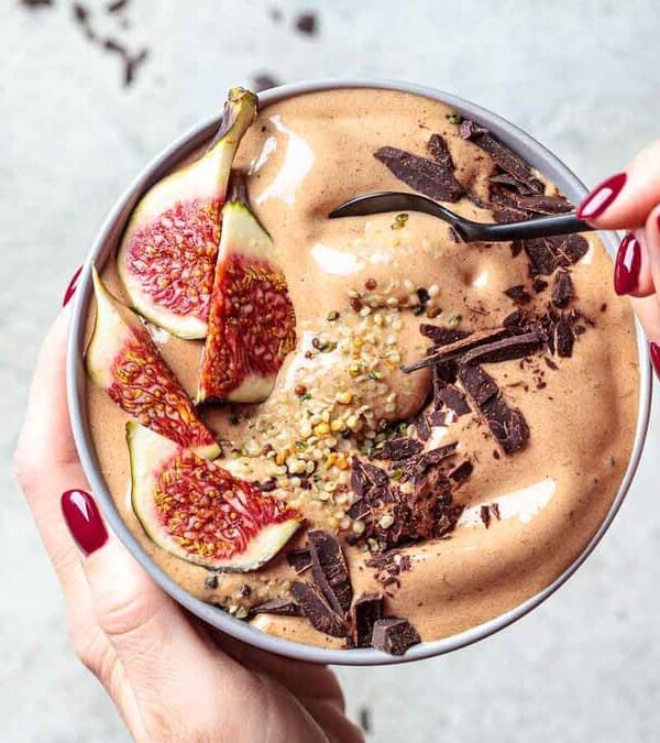 A person with red-painted nails holds a bowl of chocolate smoothie topped with sliced figs, chocolate shavings, and seeds, using a spoon to scoop from the bowl.
