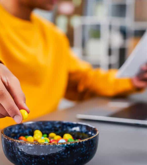 Person in yellow sweater holding a yellow candy from a blue bowl while reading a document at a desk.