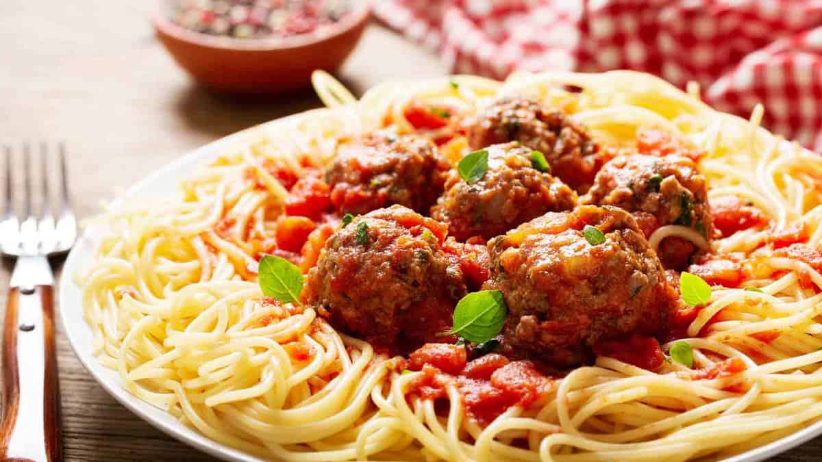 A plate of spaghetti topped with meatballs, marinara sauce, and garnished with fresh basil leaves. A red checkered cloth is in the background.