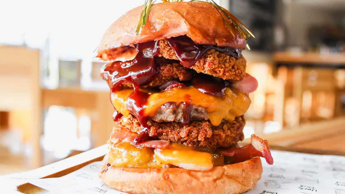 A close-up of a stacked burger with multiple layers, including fried chicken, cheese, bacon, and barbecue sauce, on a bun.