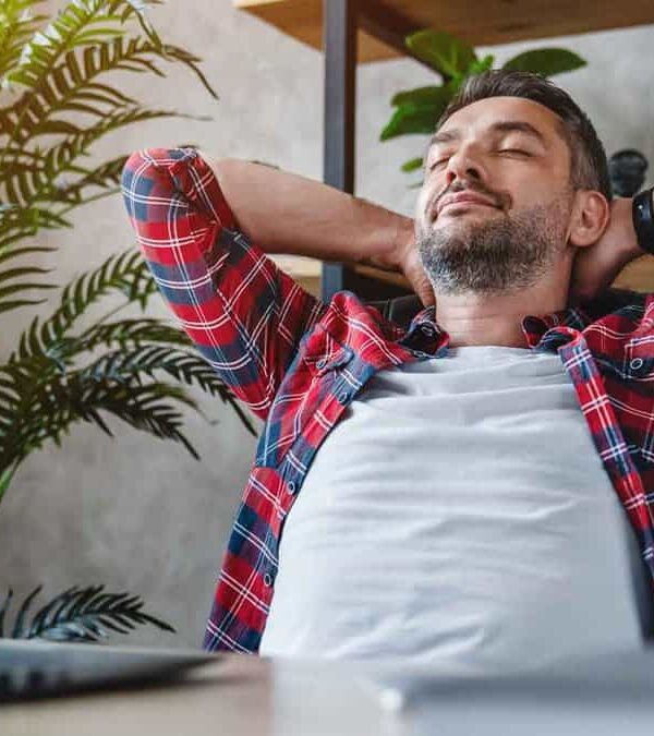 A man in a plaid shirt leans back in his chair with hands behind his head, appearing relaxed. A laptop and coffee cup are on the table in front of him, and a plant and bookshelf are in the background.