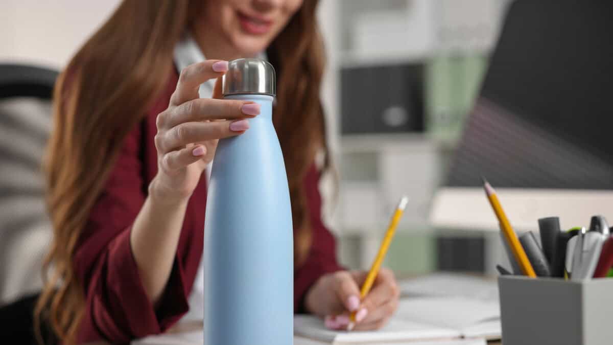 A person in a red blazer sits at a desk, holding a light blue insulated water bottle with one hand and writing in a notebook with the other. Office supplies are visible.