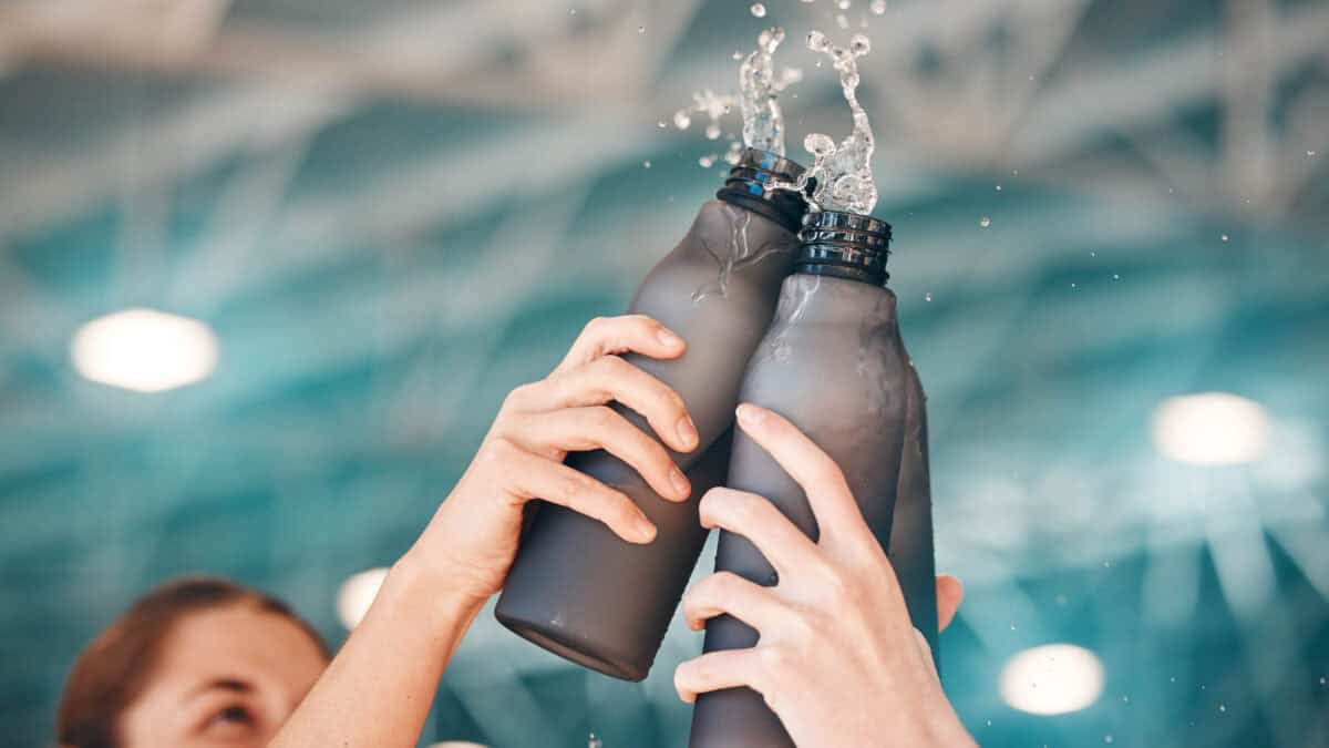 Two hands clink black reusable water bottles, causing water to splash out. The background is blurred, featuring a blue and white indoor setting.