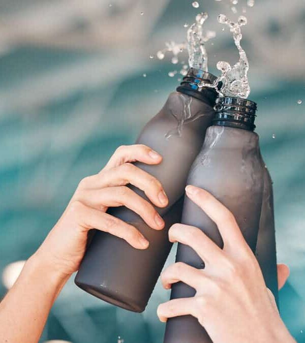 Two hands clink black reusable water bottles, causing water to splash out. The background is blurred, featuring a blue and white indoor setting.