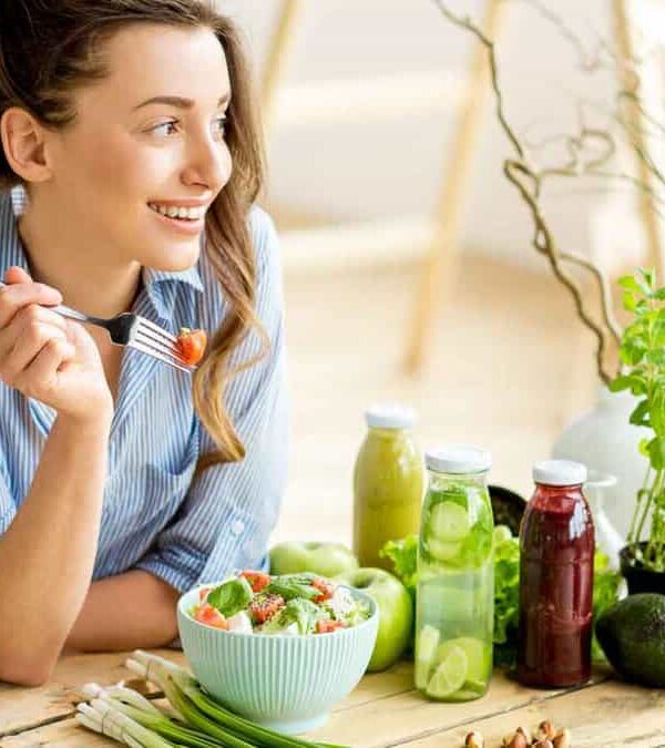 A woman sits at a table, smiling while holding a fork with a piece of food. The table has bottles of juice, herbs, and a salad.