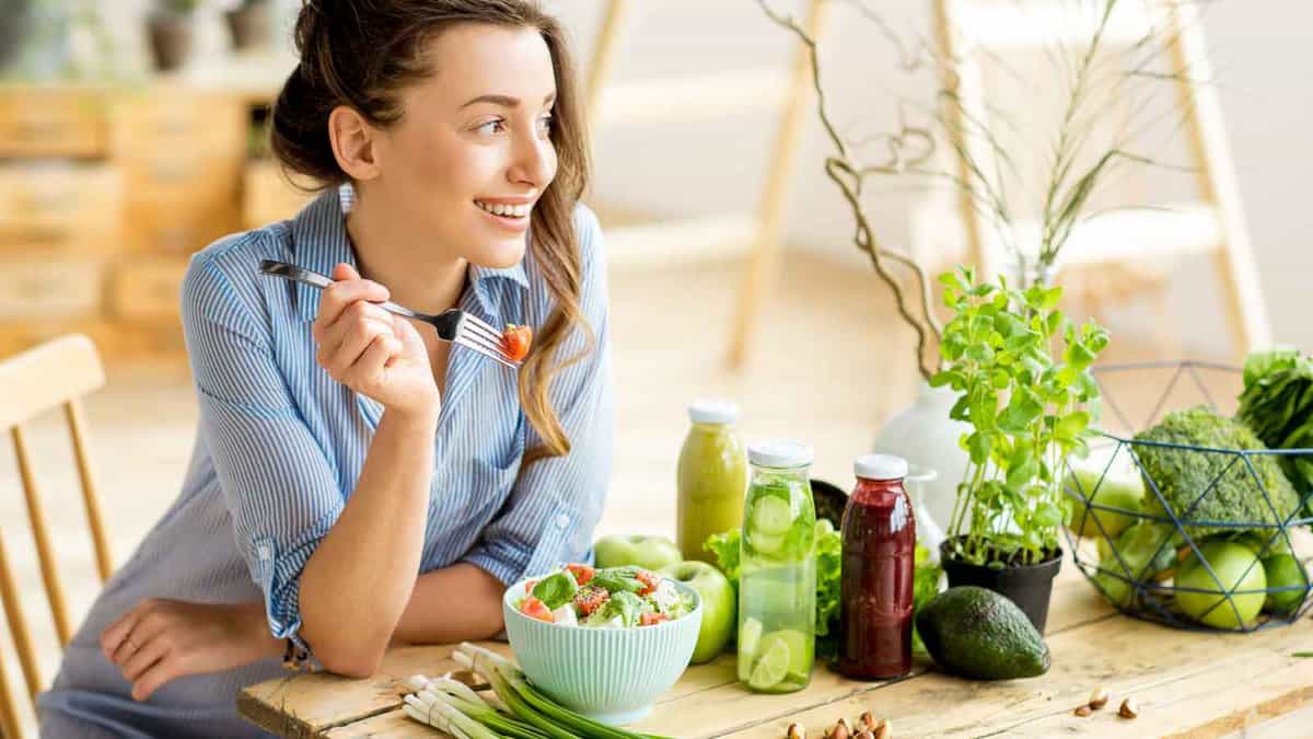 A woman sits at a table, smiling while holding a fork with a piece of food. The table has bottles of juice, herbs, and a salad.