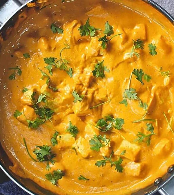 A pan filled with creamy orange-colored chicken curry, garnished with fresh cilantro, placed on a dark blue cloth on a light gray surface.