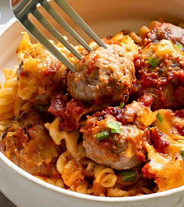A bowl of pasta mixed with tomato sauce, meatballs, and green garnishes, with a fork picking up a meatball.
