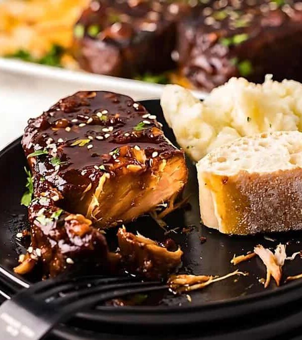 A plate with a cooked meat slab covered in sauce, a piece of bread, and mashed potatoes. Another plate of meat is in the background on a marble surface.