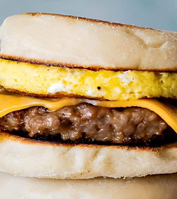 A breakfast sandwich with an English muffin, egg patty, cheddar cheese, and sausage patty, placed on a wooden surface.