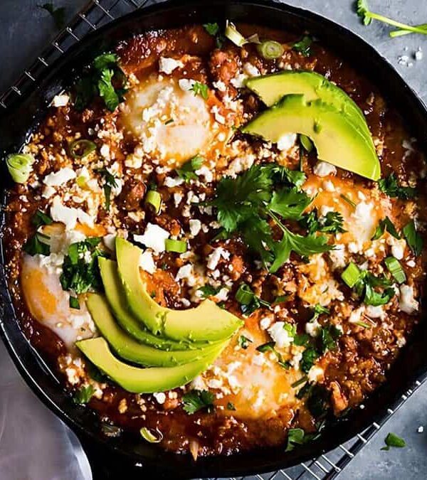 A skillet with shakshuka topped with avocado slices, crumbled cheese, and fresh cilantro. Garnished with scallions on the side and accompanied by a piece of bread and a napkin.
