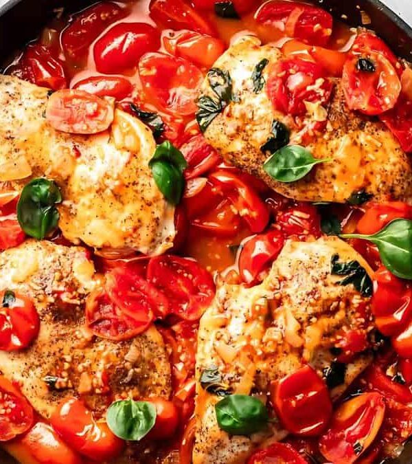 A skillet filled with cooked chicken breasts topped with fresh tomato slices, basil leaves, and a tomato-based sauce.