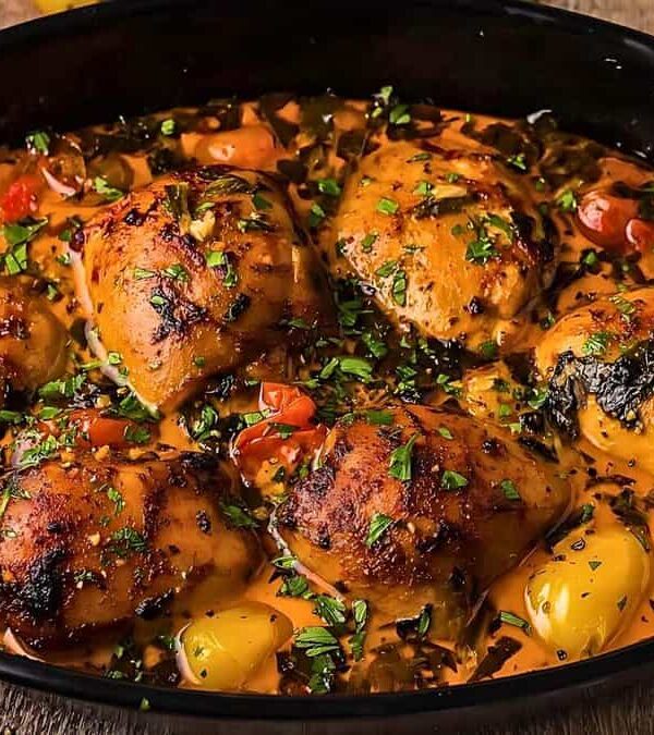 A black skillet filled with cooked chicken thighs, cherry tomatoes, and chopped herbs in a savory sauce.
