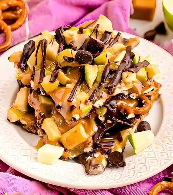 A plate of caramel apple nachos with pretzels, apple slices, chocolate chips, and caramel drizzle, placed on a purple cloth with scattered pretzels and apple slices around.
