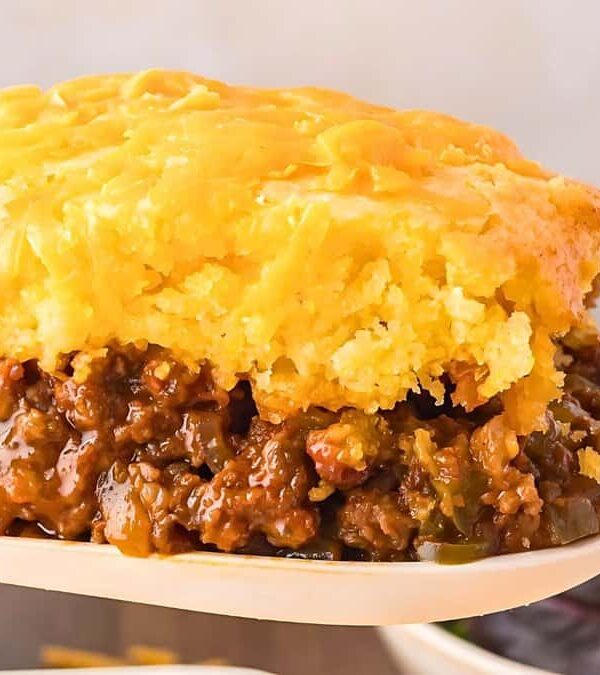 A close-up of a slice of cornbread-topped casserole, with a layer of seasoned ground meat and vegetables, being served on a spatula.