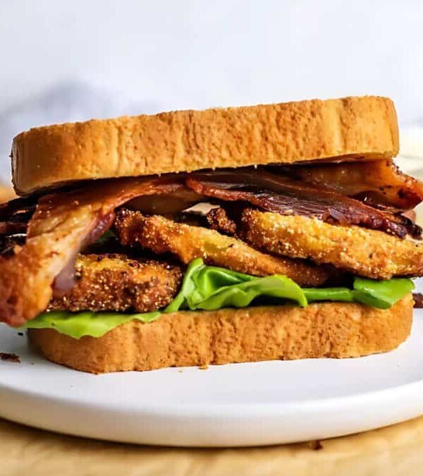 A sandwich with crispy bread, lettuce, bacon, and fried green tomatoes is placed on a white plate, with cucumber slices and a bowl of sauce in the background.