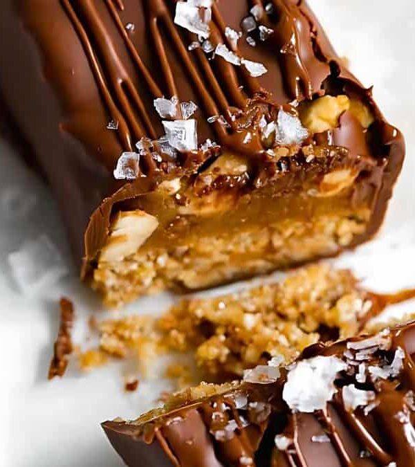 A close-up of a chocolate-covered caramel and nut candy bar, partially broken to show the inside texture, sprinkled with sea salt, reminiscent of those found in homemade snack bar recipes.