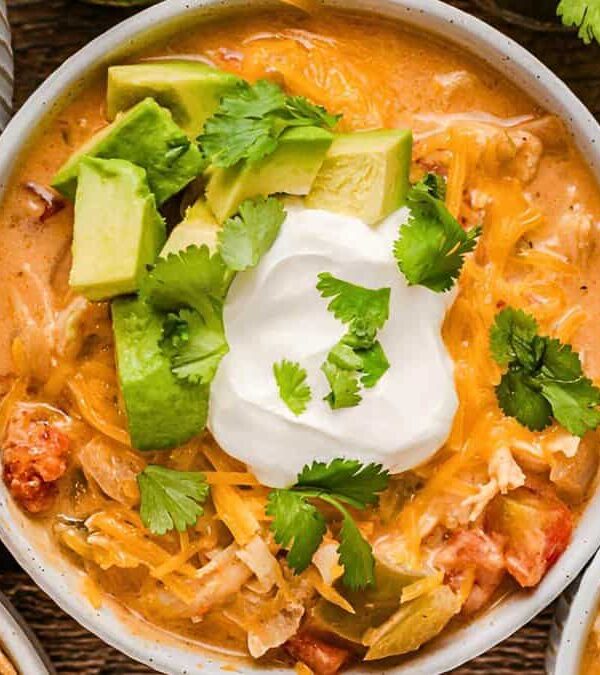 A bowl of chicken tortilla soup topped with diced avocado, shredded cheese, a dollop of sour cream, and cilantro. Surrounding it are bowls of sour cream and tortilla chips.