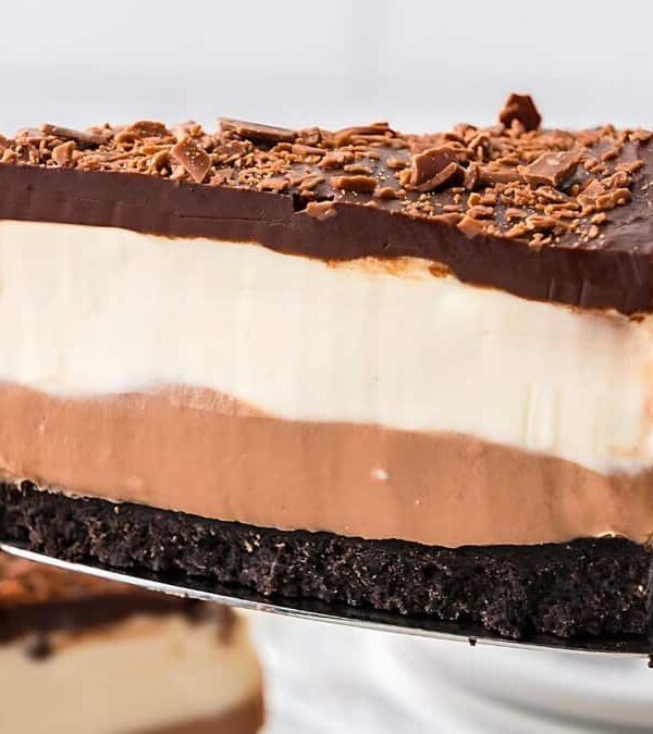 A slice of triple-layered chocolate cheesecake with a base of chocolate crust, a middle layer of chocolate mousse, and a top layer of chocolate ganache, garnished with chocolate shavings.