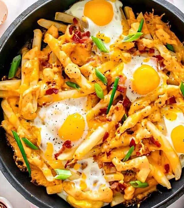 A skillet filled with cheese-covered fries, topped with sunny-side-up eggs, bacon bits, and chopped green onions. Lime halves, dipping sauce, and broken eggshells are on the side.
