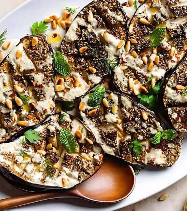 A white platter holds roasted eggplant halves topped with tahini sauce, pine nuts, and fresh herbs. A wooden spoon rests alongside. Fresh herbs are scattered around the dish on the table.