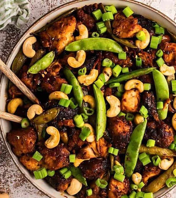 A bowl of stir-fried chicken with cashews, snap peas, green onions, and a savory sauce. Chopsticks rest on the side of the bowl.