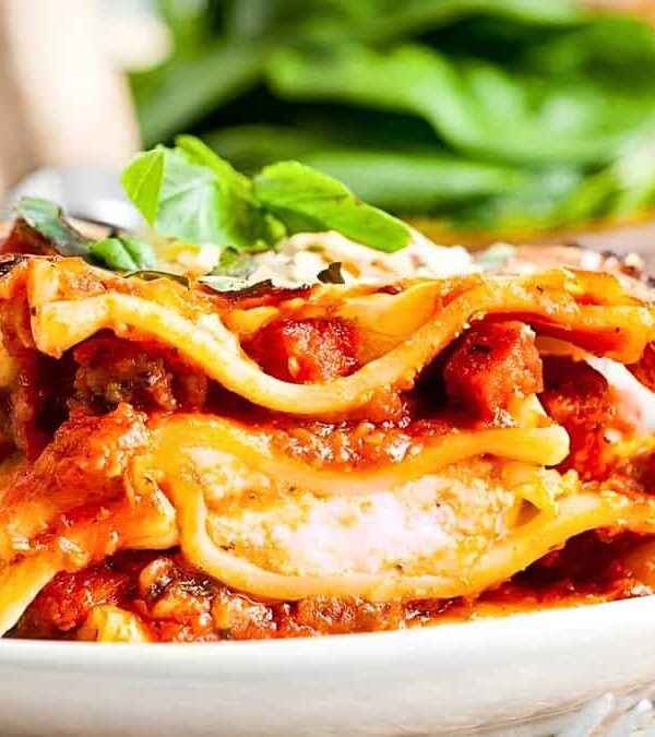 A serving of lasagna on a white plate, showing layers of pasta, meat, cheese, and sauce, topped with a basil leaf.