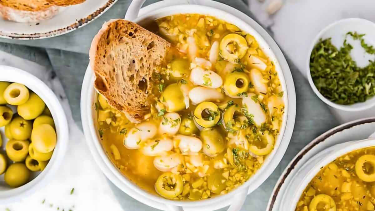 A bowl of soup with white beans, green olives, herbs, and a slice of bread, surrounded by a small bowl of chopped herbs and another bowl of green olives.