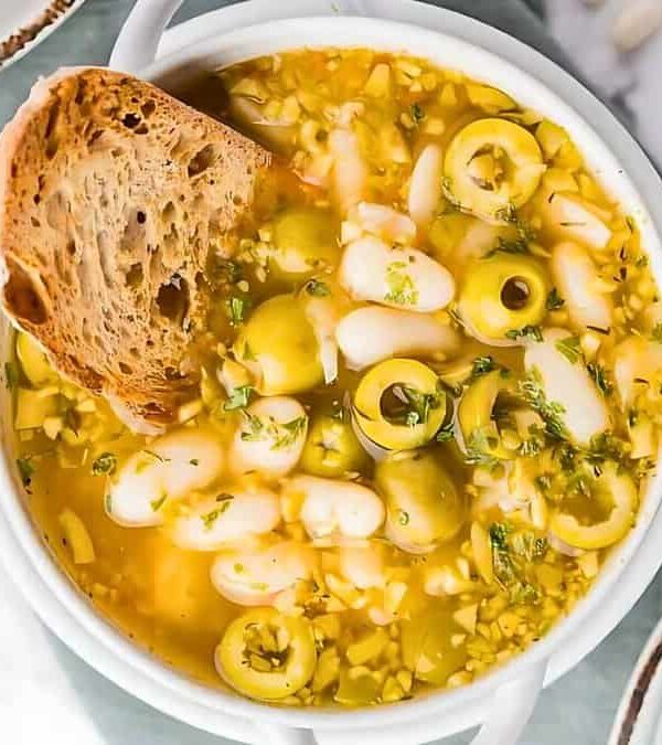 A bowl of soup with white beans, green olives, herbs, and a slice of bread, surrounded by a small bowl of chopped herbs and another bowl of green olives.