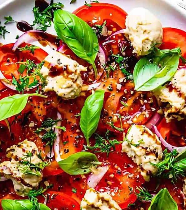 A plate of sliced tomatoes, red onions, fresh basil leaves, and dollops of hummus, drizzled with balsamic vinaigrette, and garnished with chopped herbs. Forks and a napkin are placed nearby.