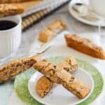 A plate with two pieces of biscotti on a green and white napkin. Additional biscotti, a cup of coffee, and a tea set are in the background.