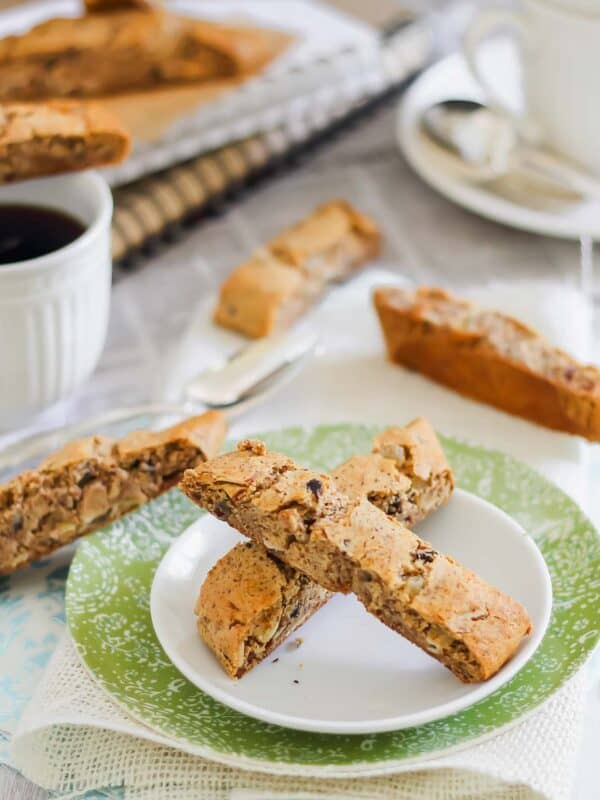 A plate with two pieces of biscotti on a green and white napkin. Additional biscotti, a cup of coffee, and a tea set are in the background.