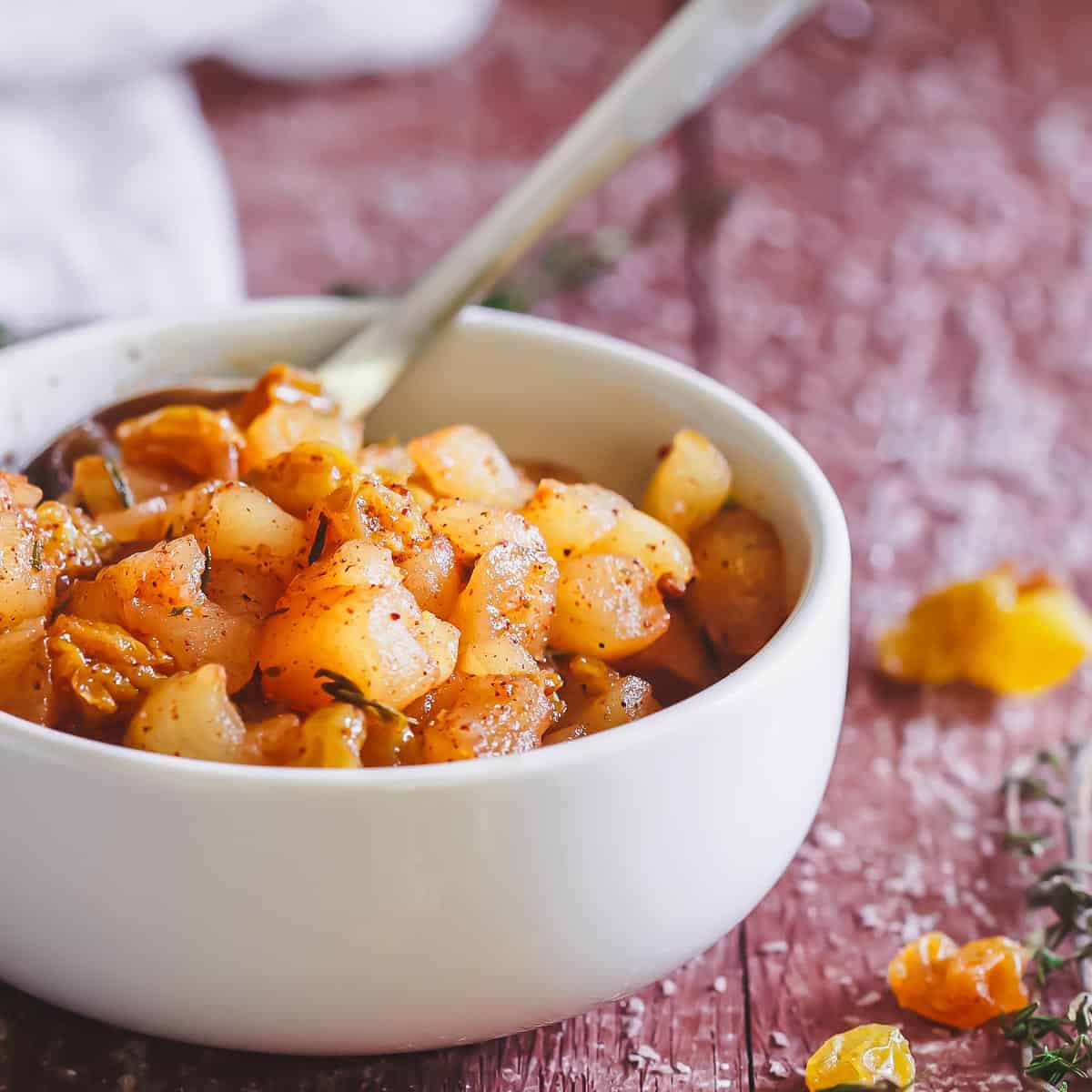 A white bowl filled with cooked, seasoned diced apples, raisins and herbs, sitting on a rustic wooden surface with a spoon inside.