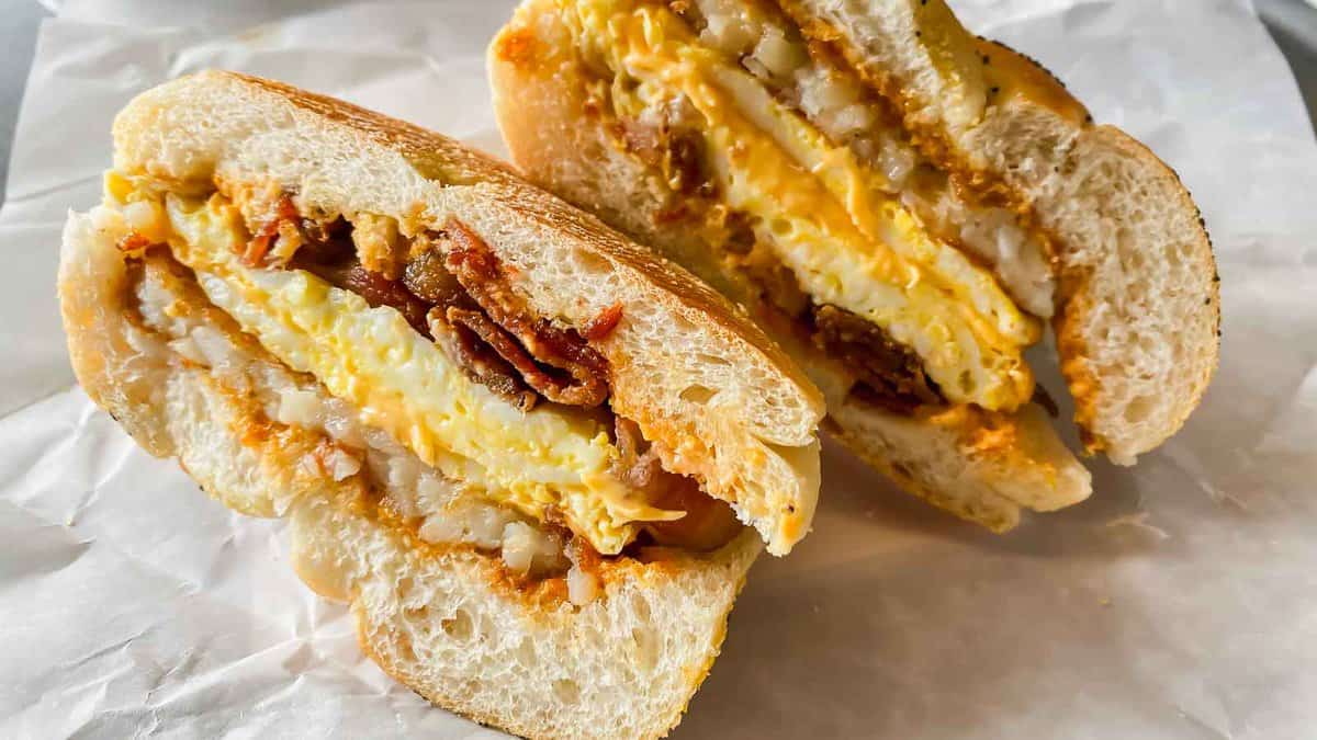 A breakfast sandwich cut in half, filled with egg, bacon, cheese, and hash browns, served on a white paper.