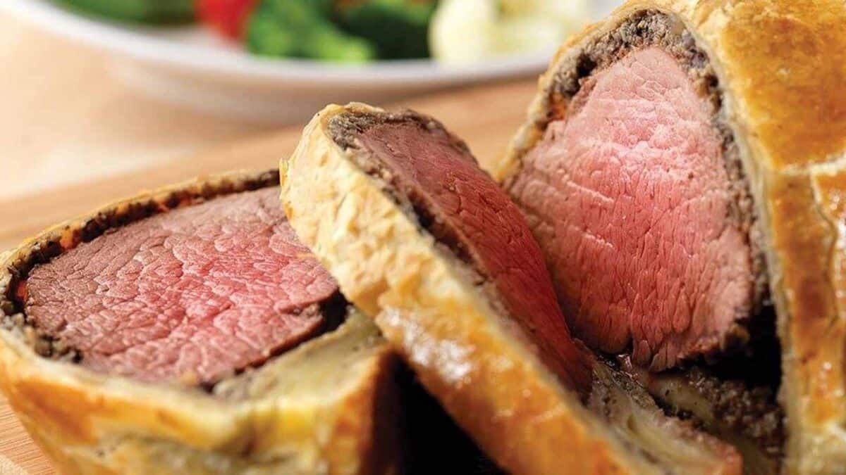 Close-up of sliced Beef Wellington showing medium-rare beef wrapped in puff pastry, with a plate of vegetables in the background.