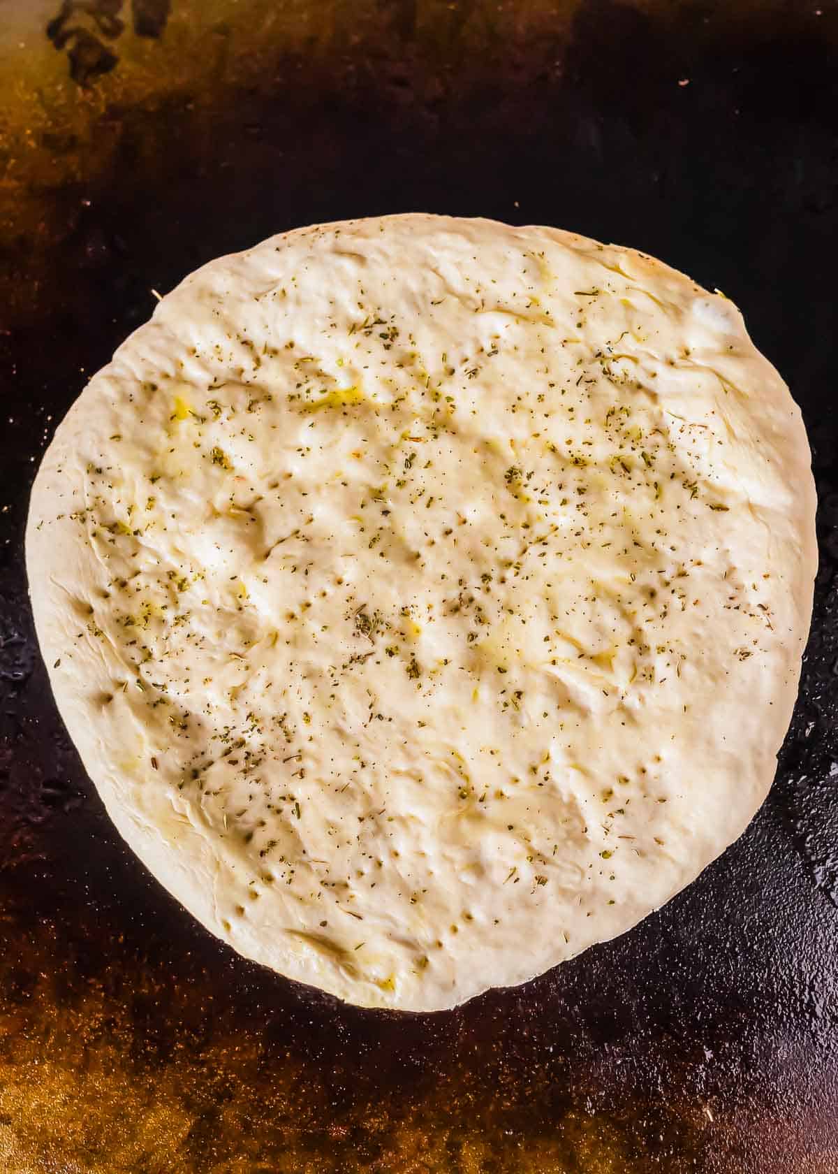A round, uncooked pizza dough topped with herbs and seasoning on a griddle.