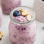 A glass jar filled with blueberry chia pudding topped with sliced almonds and a whole blueberry. Other jars of similar pudding are visible in the background.