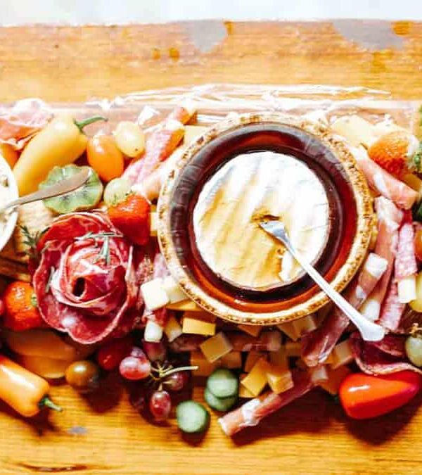 A long wooden board is topped with a variety of fresh fruits, vegetables, cheeses, dips, cured meats, and breadsticks. In the center, there is a dish of dip with a spoon.