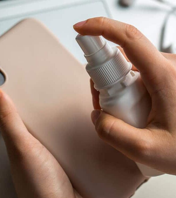 Person cleaning a smartphone case with a spray bottle near a laptop and earphones on a table.