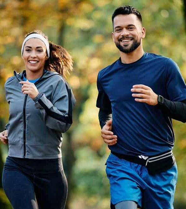 Two people are jogging together in a forested area while smiling, one wearing a gray jacket and the other in a blue shirt.
