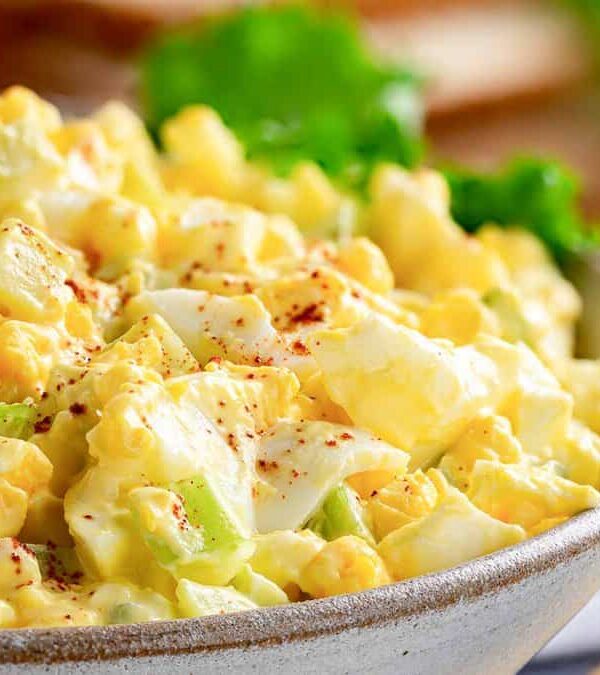 A close-up of a bowl of potato salad, featuring chunks of potatoes, boiled eggs, and celery, sprinkled with paprika. Green lettuce is visible in the background.