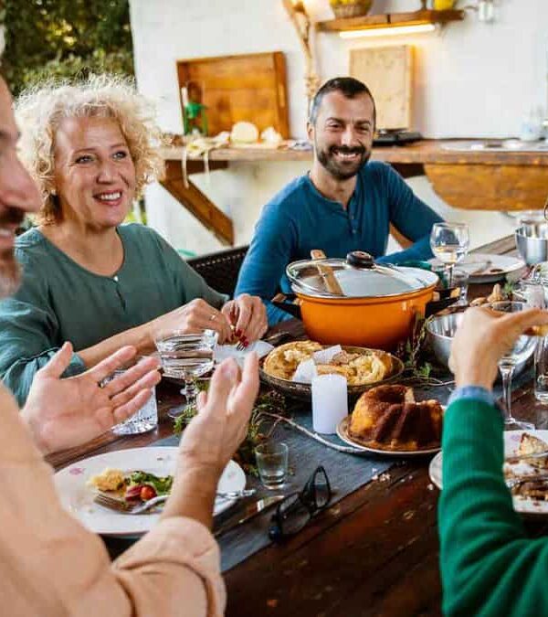 A group of people sit around a wooden table outdoors, sharing a meal and smiling. Various dishes, utensils, and glasses are spread across the table.