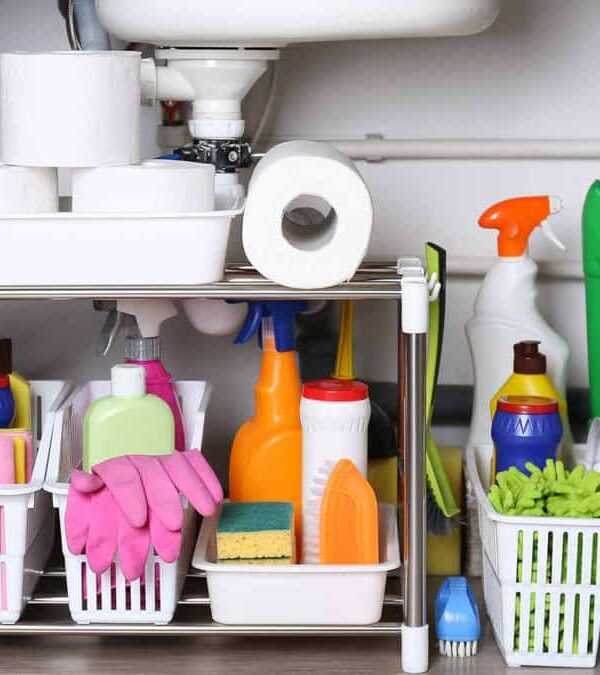 A cabinet under a sink organized with cleaning supplies, including sponges, gloves, spray bottles, brushes, and paper towels. Different items are stored in baskets and on shelves.