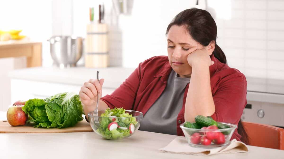 A woman in a red shirt and brown jacket looks unenthusiastically at a salad in a bowl with a fork, with vegetables including lettuce, radishes, cucumber, and an apple on the kitchen counter.