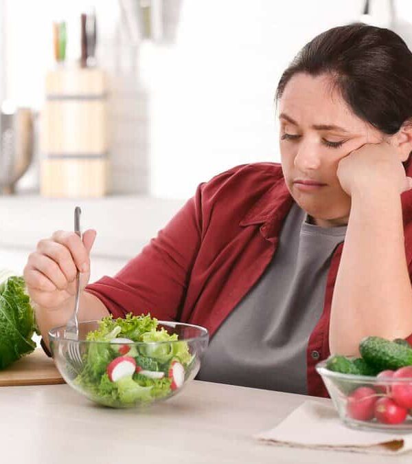 A woman in a red shirt and brown jacket looks unenthusiastically at a salad in a bowl with a fork, with vegetables including lettuce, radishes, cucumber, and an apple on the kitchen counter.