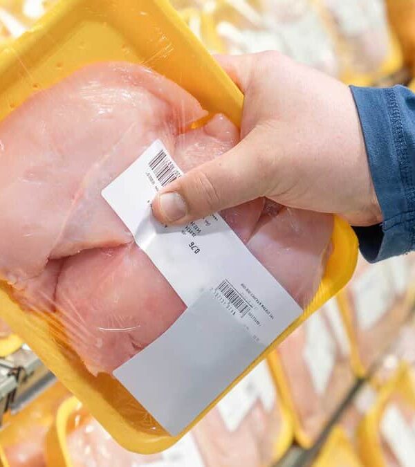 A person in a blue shirt is holding a packaged tray of raw chicken breast in a supermarket aisle.