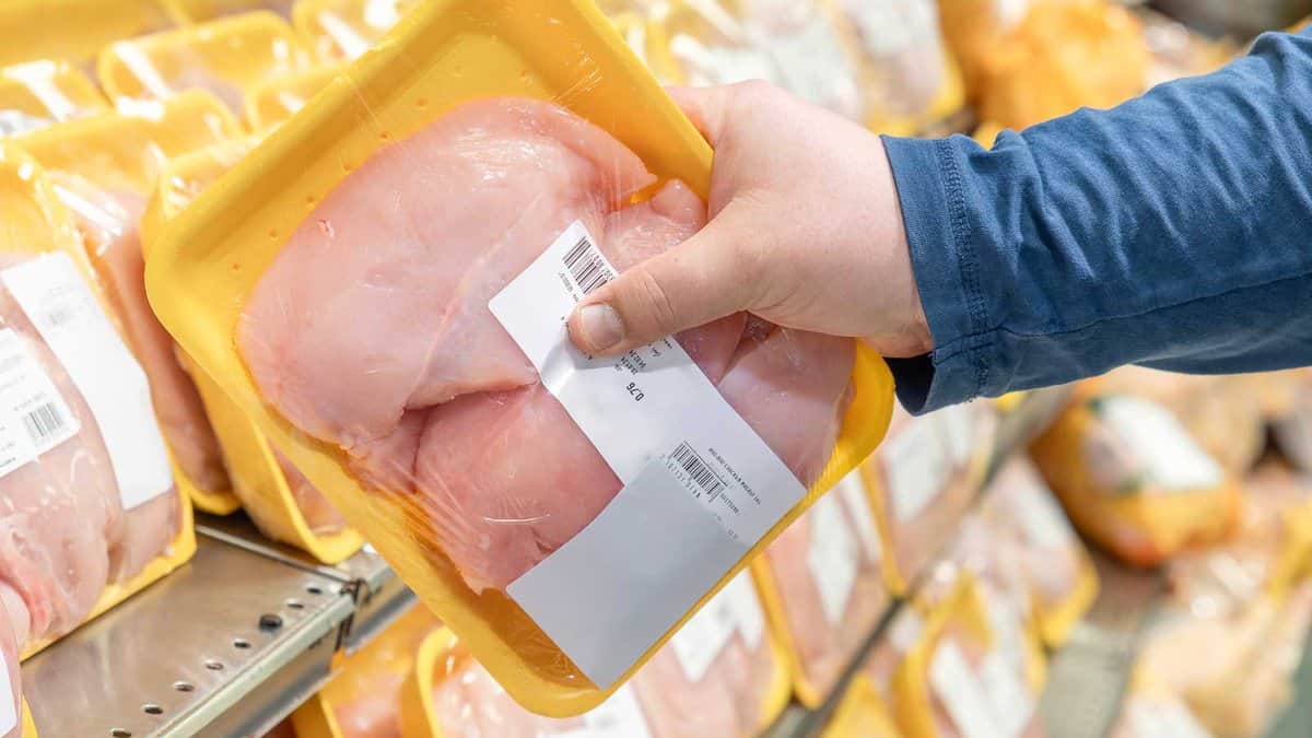 A person in a blue shirt is holding a packaged tray of raw chicken breast in a supermarket aisle.