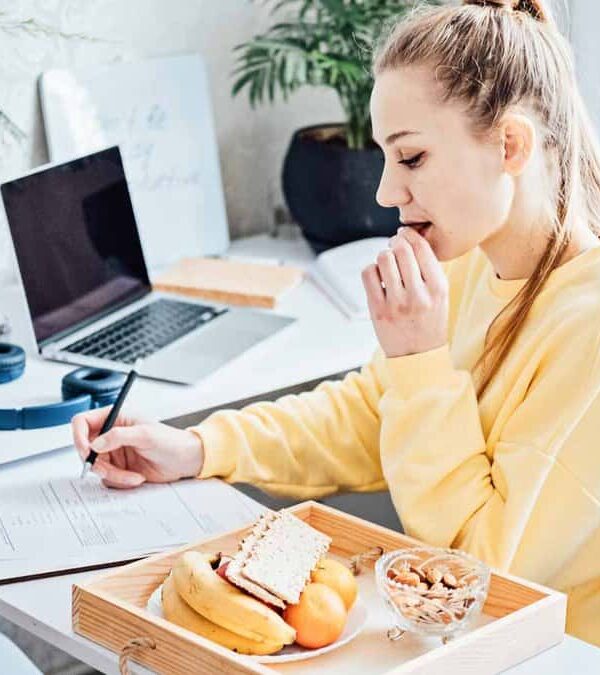 A woman in a yellow sweater works at a desk with papers, a laptop, and a tablet. She is eating snacks from a tray that includes fruit and nuts. The desk has plants and headphones.